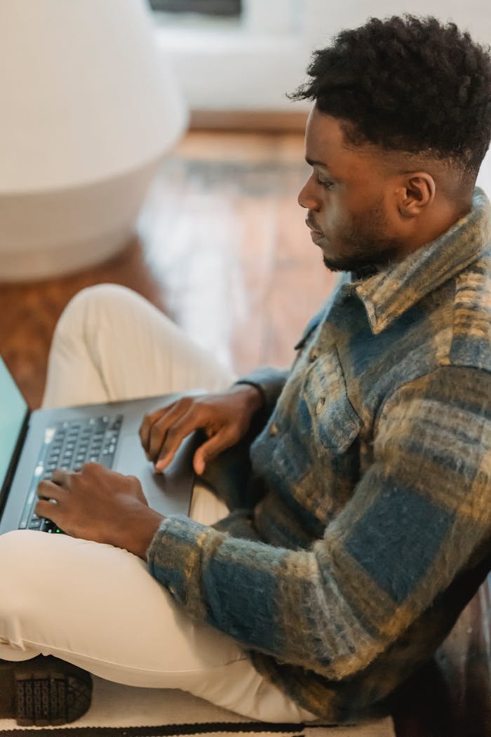 about-02 Side view of concentrated African American guy sitting on parquet and browsing laptop while working remotely from home